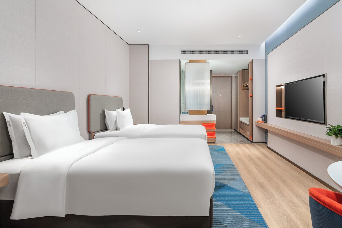 Quarto de hotel moderno com duas camas, televisão, banheiro e um espelho grande.