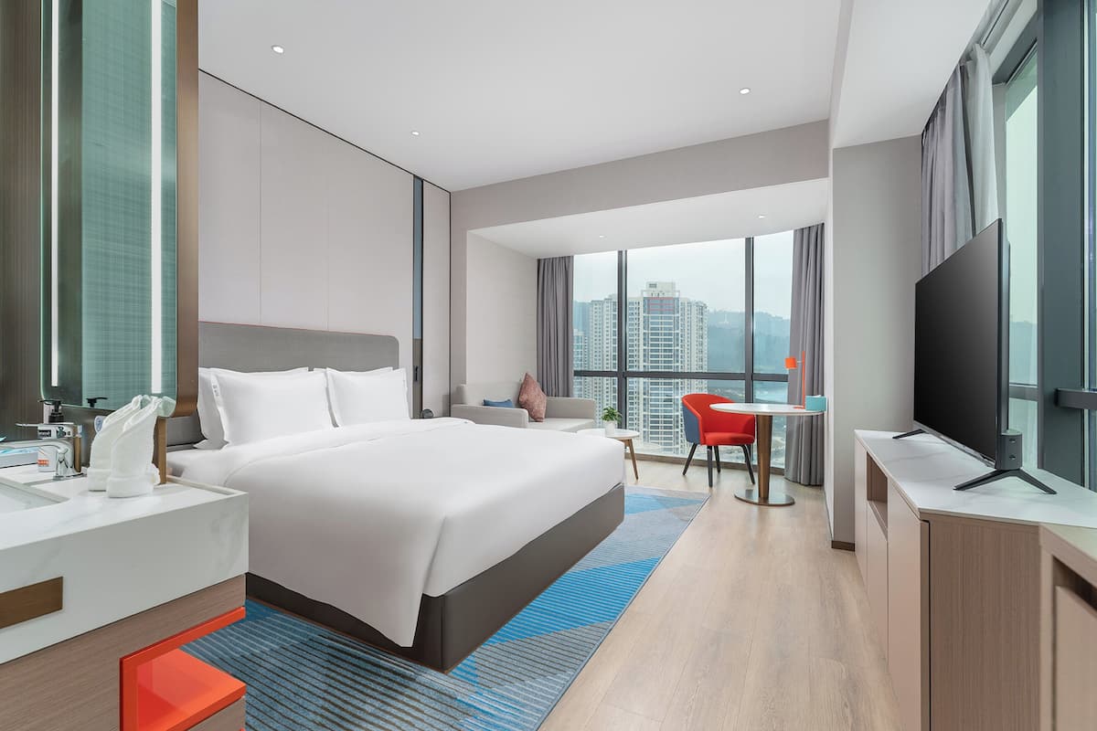 A modern hotel room with a large bed, a desk, a TV, and a view of the city.
