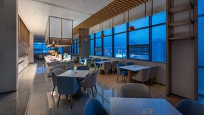 A modern dining area with tables and chairs, large windows offering a city view, and a kitchen area with a bar. - Holiday Inn Express Bazhong Center by IHG (Bazhong)
