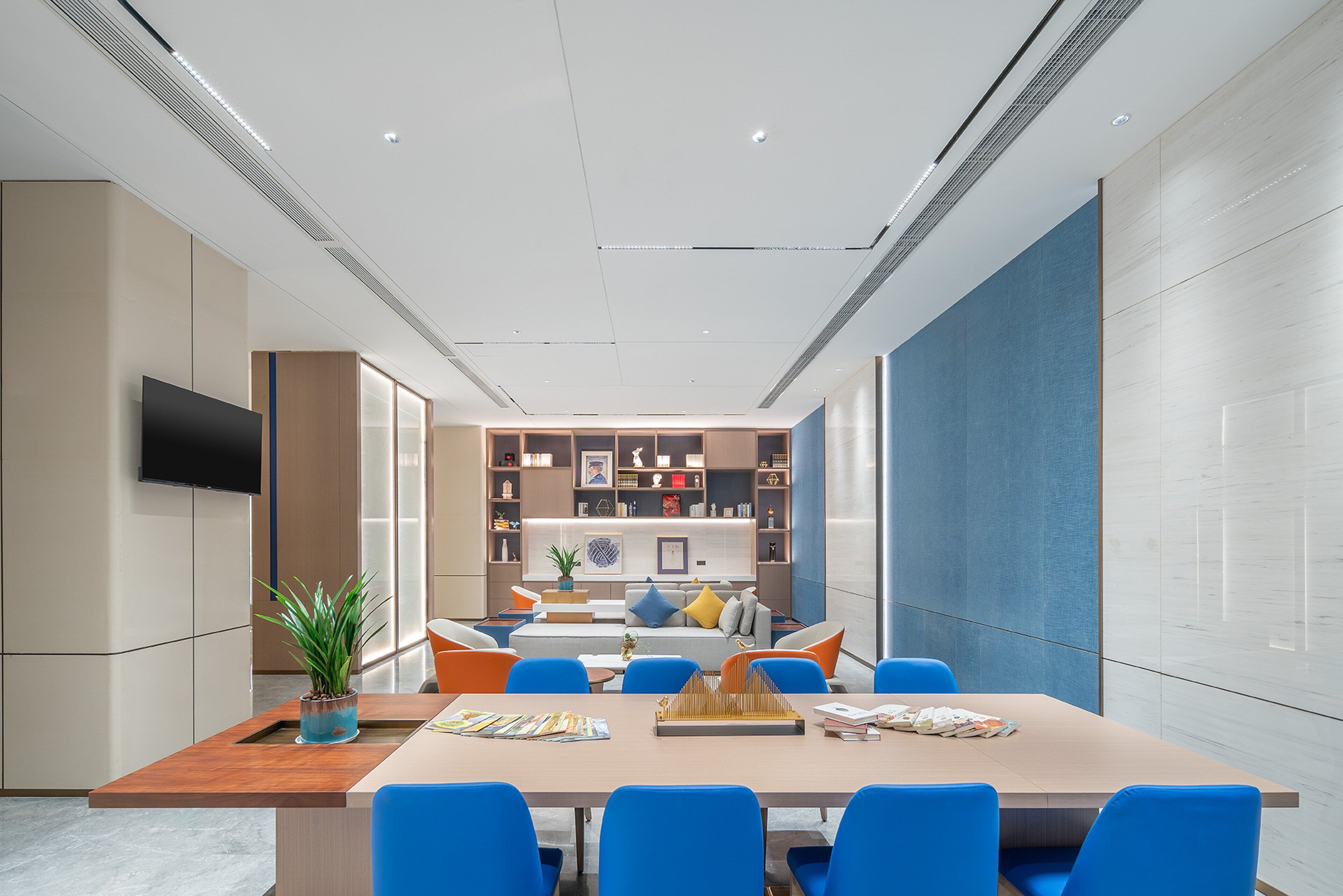A modern meeting room with a long table, blue chairs, and a television on the wall.