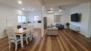 A modern kitchen with white cabinetry, a dining area with white chairs, a sofa, and a flat-screen TV mounted on the wall. - Unwind in Comfort, A Perfect Getaway Awaits Centrally Located Near All Amenities (Southport)