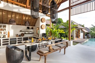 Uma área moderna de refeições ao ar livre com um banco de madeira, luminárias de vime pendentes e vista para a piscina.