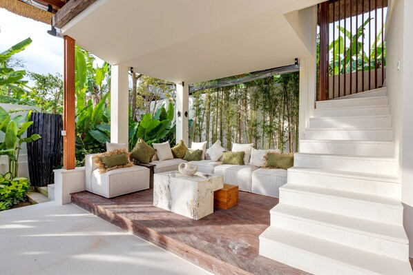 A covered outdoor seating area with white sofas and a marble coffee table, surrounded by lush greenery. - Villa Chempaka by Alfred in Bali (Kerobokan)