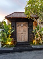 Uma porta de madeira tradicional com esculturas detalhadas, ladeada por plantas tropicais exuberantes e um caminho de pedra que leva à entrada.