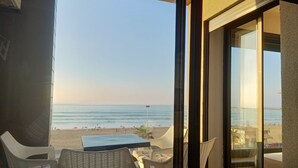 Un balcón con vista a la playa, sillas de mimbre, una mesa y una puerta de vidrio que da a un espacio interior.
