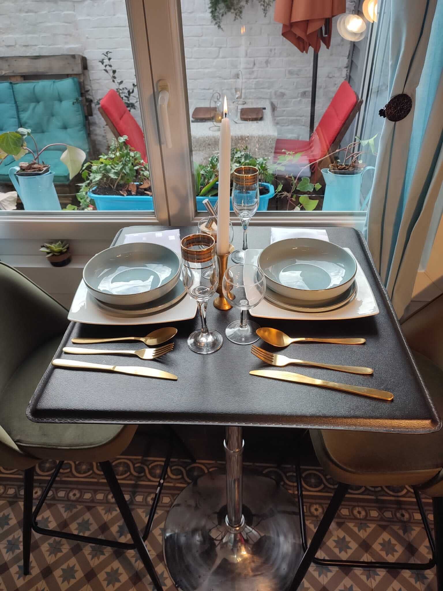 A table set for two with plates, cutlery, and a candle, viewed through a window overlooking a brick wall and outdoor seating area.