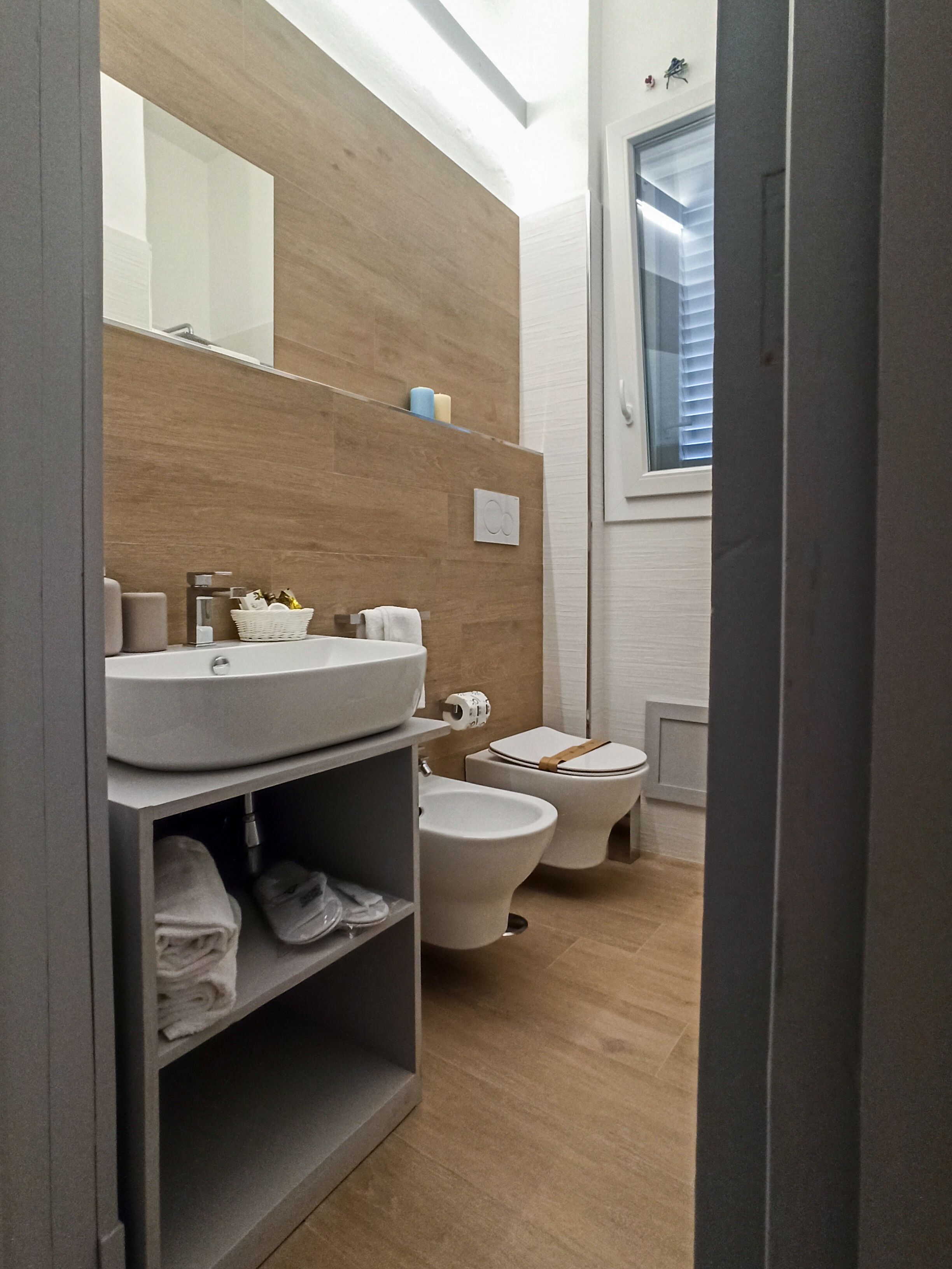 Une salle de bain moderne avec un grand miroir, un lavabo, des toilettes et un bidet. On y trouve une étagère avec des serviettes et une fenêtre avec des stores.