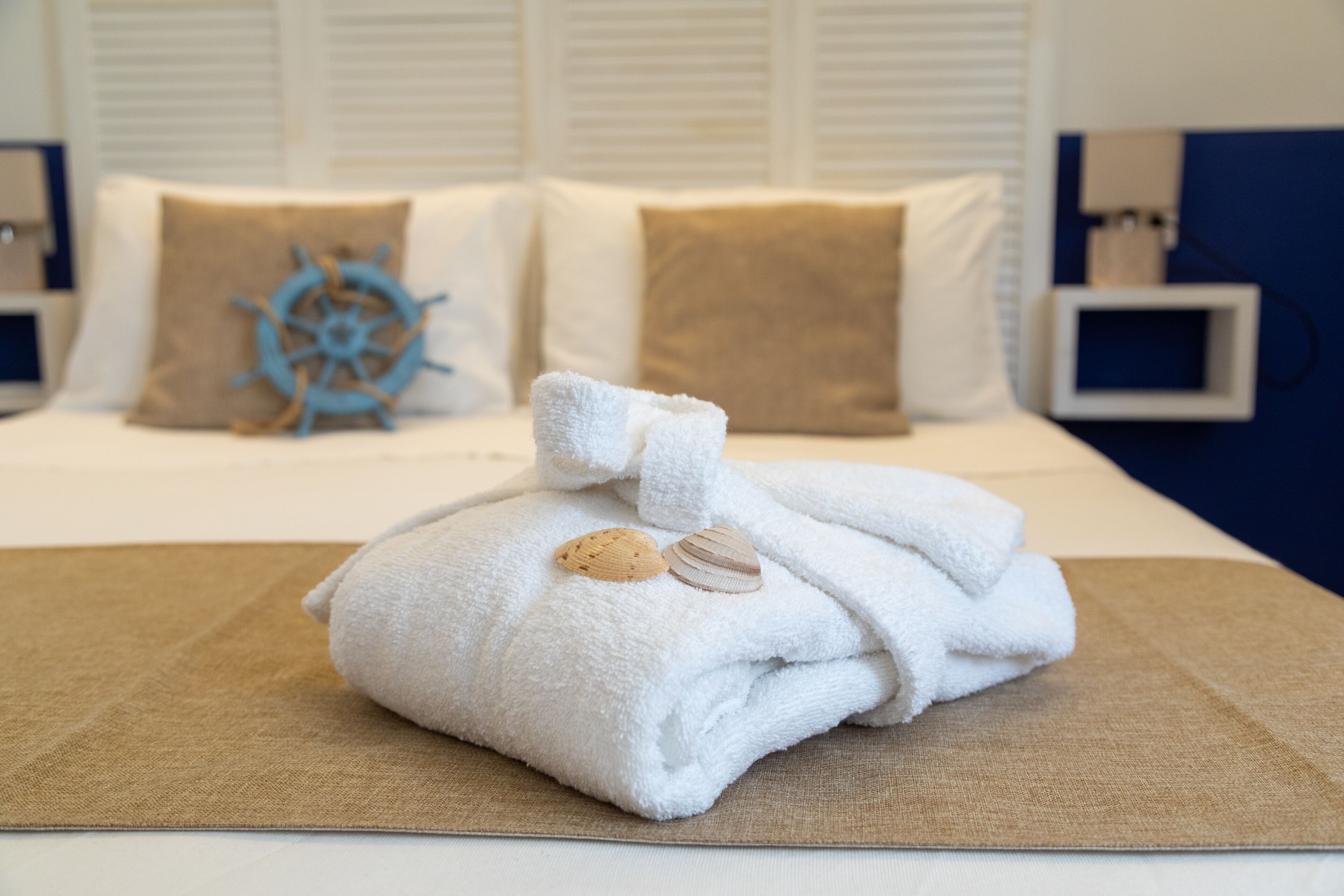 Una cama bien hecha con una toalla y conchas marinas, una decoración con timón de barco y una mesita de noche con una lámpara.