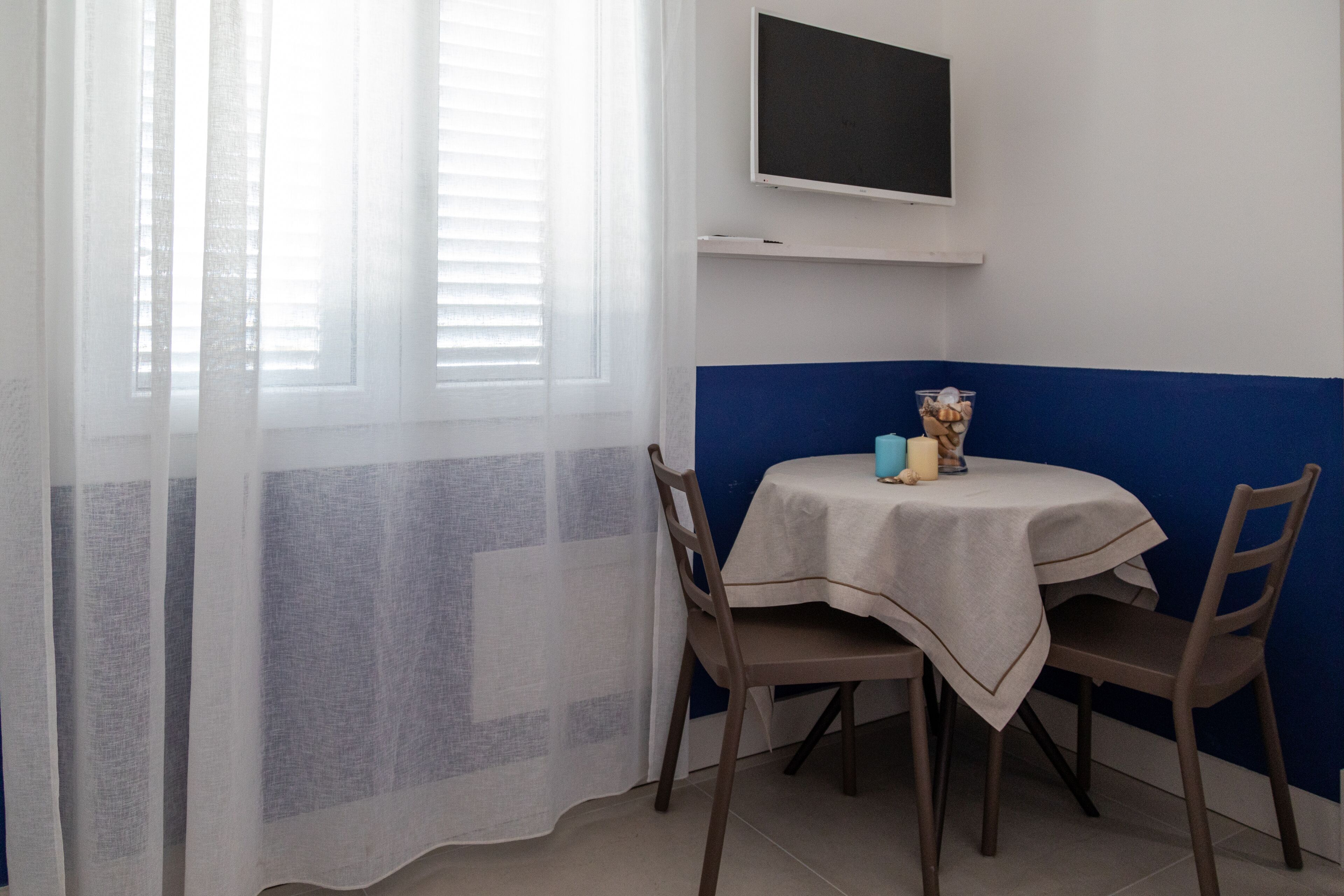 Un pequeño comedor con una mesa, sillas, un televisor montado en la pared y una ventana con cortinas traslúcidas.