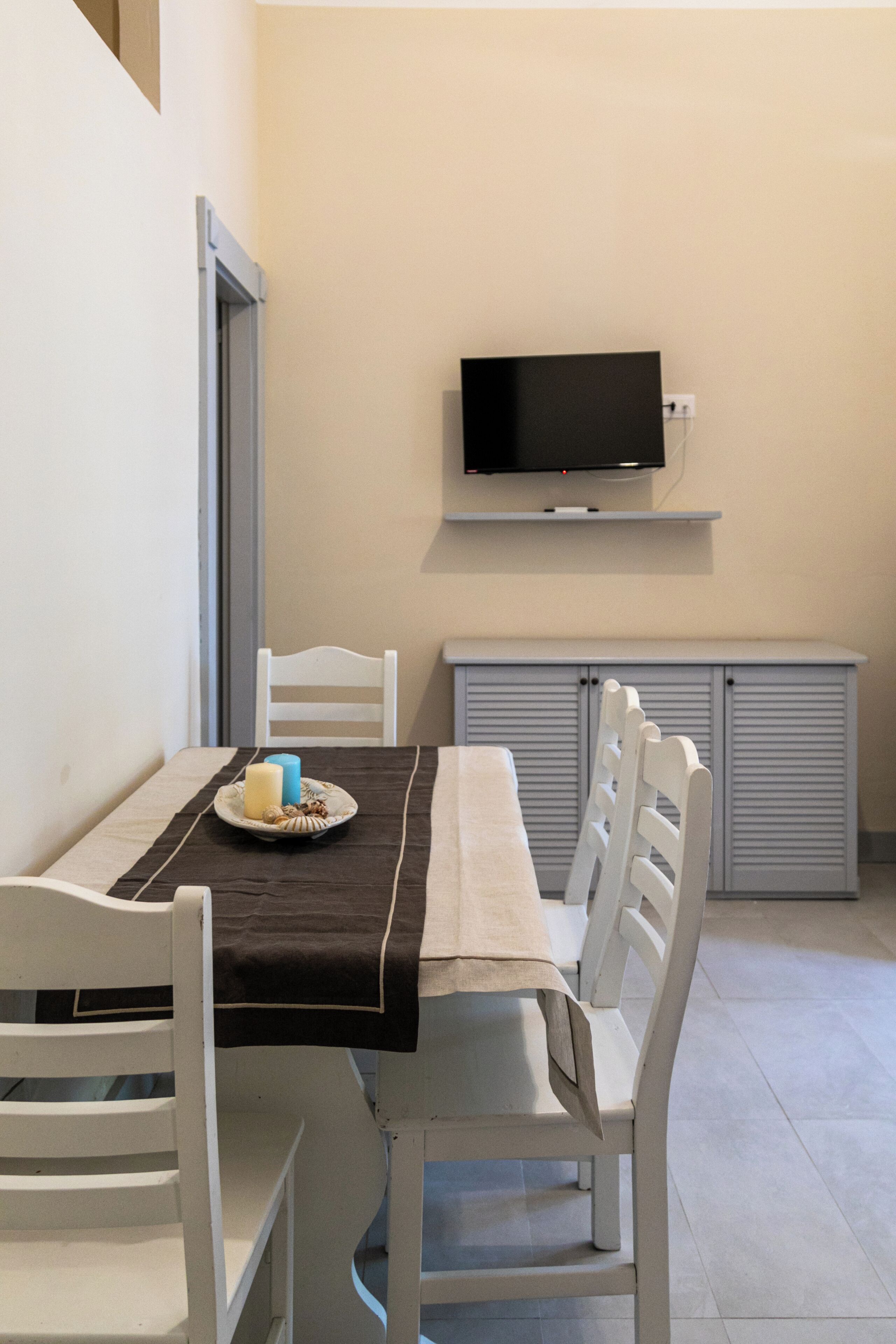 A dining area with a table set for two, a television mounted on the wall, and a white chair.