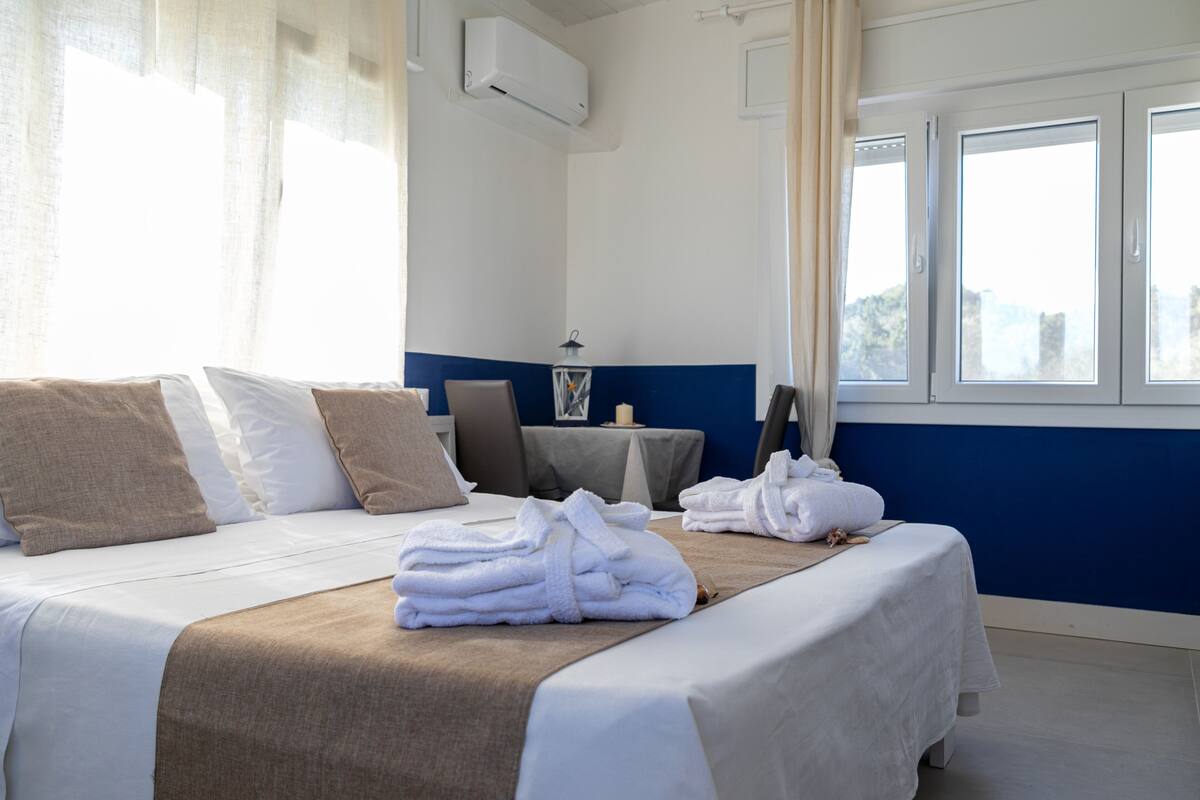 A hotel room with a bed, pillows, a bedside table with a lamp, and a window with curtains.