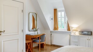 Une chambre avec un plafond en pente, un bureau avec une chaise, un lit, une fenêtre avec des rideaux, un miroir et une table de chevet avec une lampe et une radio.