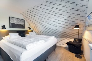 Ein ordentlich gemachtes Bett mit weißen Leinen, einem schwarzen Kopfteil und zwei Kissen mit Aufdruck. Eine schwarze Stehlampe und ein schwarzer Sitzsack sind ebenfalls vorhanden. Der Raum hat Tapeten mit Fischmotiven und ein gerahmtes Bild an der Wand.