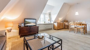 Een ruime loft met hoge plafonds, een eethoek met houten stoelen en een tafel, een glazen salontafel, een televisie op een houten kast, en een groot raam dat voor natuurlijk licht zorgt.