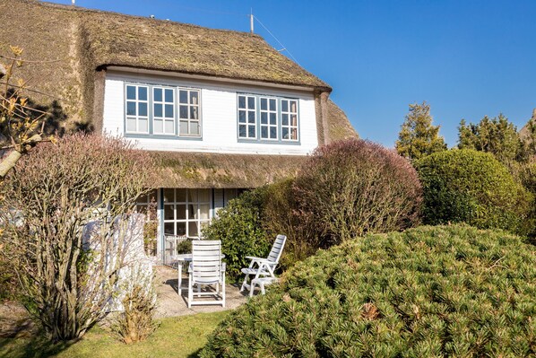 A traditional thatched-roof house with a white exterior, surrounded by a well-maintained garden with a lawn and shrubs. - Moers - Wohnen Unter Reet im Herzen von Keitum (Sylt / OT Keitum)