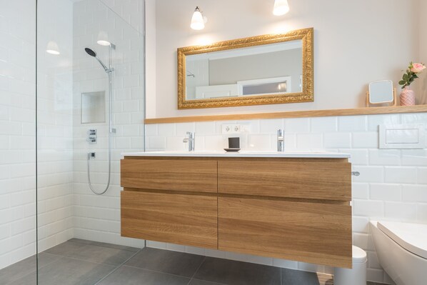 A modern bathroom with a glass shower enclosure, a wooden vanity with a white countertop, and a large mirror with a decorative frame. - Schöne Erdgeschoßwohnung auf Einer Ebene, ca. 130 m² Groß, für bis zu 4 Personen (Sylt / OT Keitum)