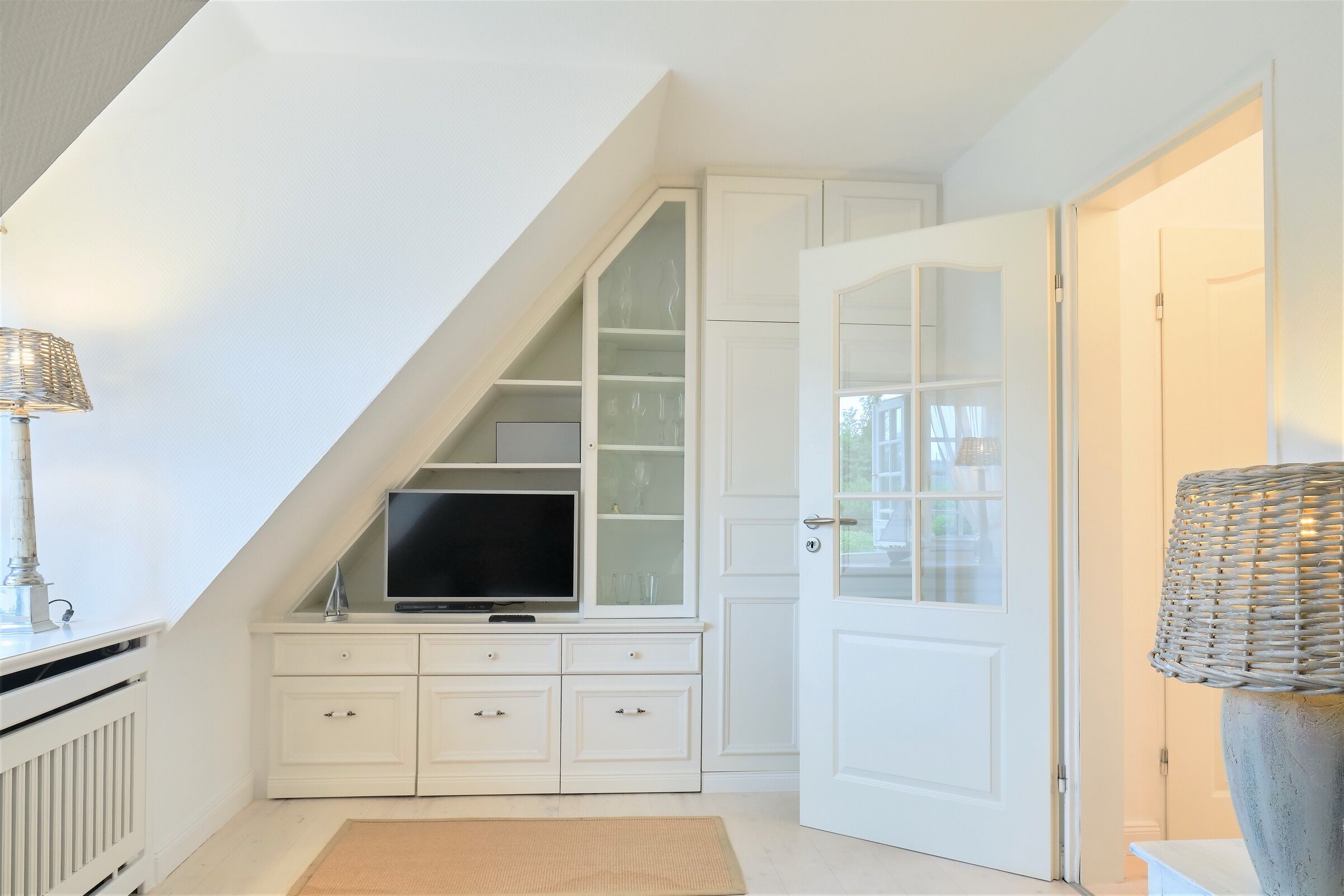 Ein Zimmer mit schrägen Wänden, weißen Schränken mit Glastüren, einem Flachbildfernseher und einer Weidenkorzlampe.