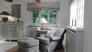 A modern living room with a grey sofa, ottoman, and a table with a lamp. There is a radiator, a window with a view of greenery, and a cabinet with decorative items. - Wohnung Über Zwei Ebenen ca. 90m² für bis Vier Personen (Sylt / OT Keitum)