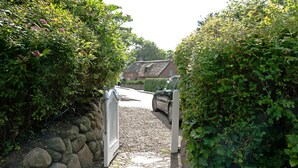 A narrow driveway leading to a house with a thatched roof, surrounded by dense greenery and a stone wall on one side. - Wohnung Über Zwei Ebenen ca. 90m² für bis Vier Personen (Sylt / OT Keitum)