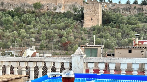 A poolside area with a glass of red wine, a bowl of blueberries, and a view of a historic castle on a hill. - Casa con Jacuzzi y Piscina con Vistas al Castillo (Corbera)