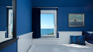 A bedroom with a view of the sea, a window with curtains, a framed painting, and blue patterned pillows.