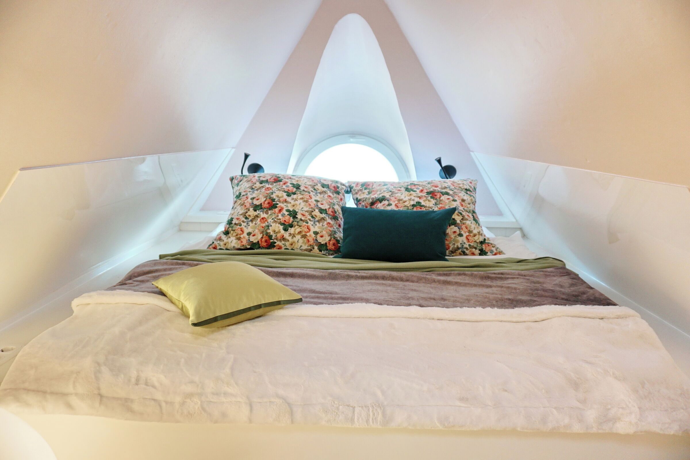 Una cama con almohadas florales, una almohada verde y una manta blanca en una habitación con un techo alto y abovedado, y un tragaluz.