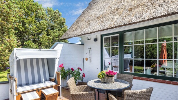 A patio area with a thatched roof, wicker furniture, and a white exterior wall. - 2-raum Appartement Unter Reet im Herzen von Keitum (Sylt / OT Keitum)