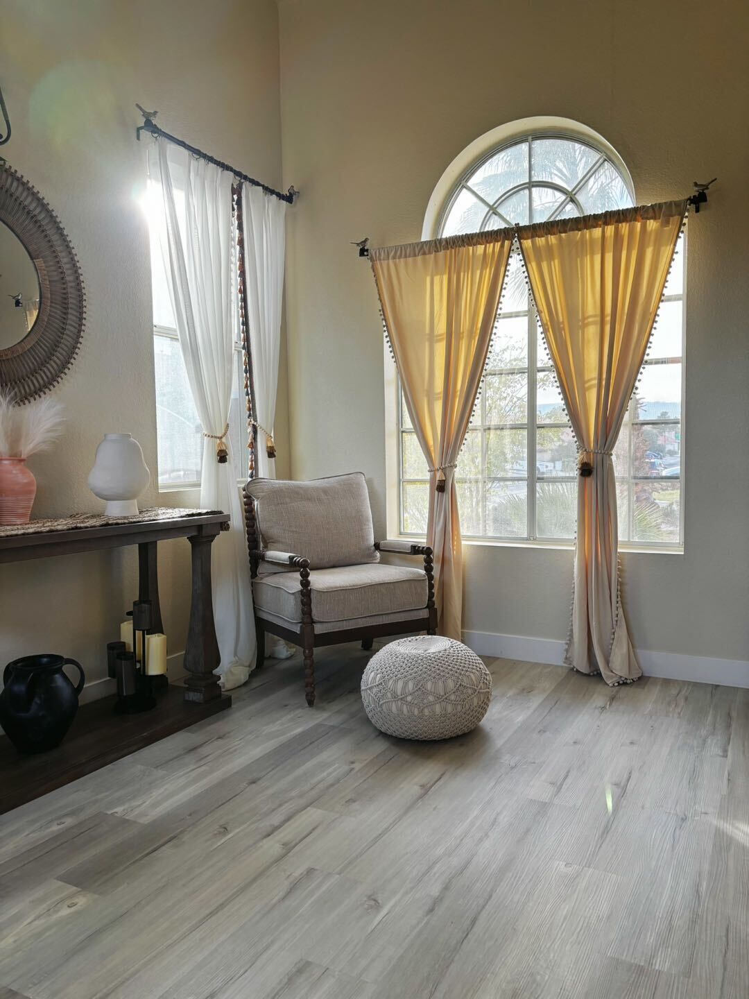 Quarto com piso de madeira, uma cadeira, um banquinho redondo, uma mesa com abajur e uma janela com cortinas.