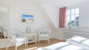 Ein Schlafzimmer mit einem Bett, zwei Korbstühlen, einem kleinen Tisch, einem Fenster mit Vorhängen und einem Wandbild.