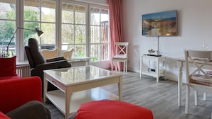Ein modernes Wohnzimmer mit einem Glastisch, einem roten Sofa, einem weißen Sessel und einem Esstisch mit weißer Tischdecke.