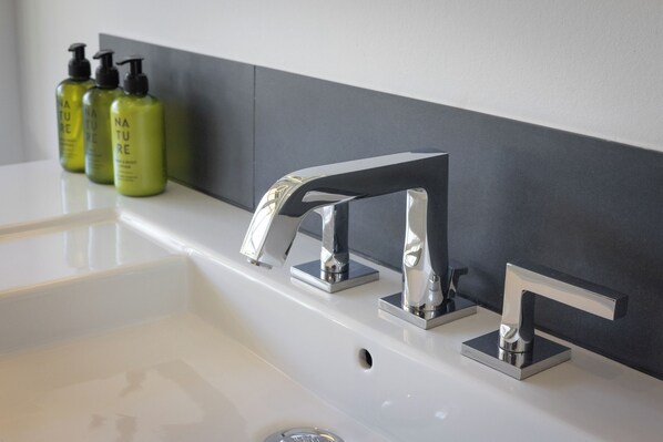 Ein modernes Badezimmer mit einem chromfarbenen Wasserhahn, einem weißen Waschbecken und drei grünen Flaschen Nature Shampoo.