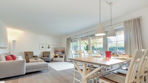 Un grand espace de vie comprenant un canapé, une table à manger et des chaises. Au fond, on trouve une cuisine avec une fenêtre donnant sur un jardin.