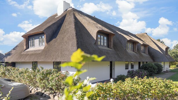 Une maison à toit de chaume, aux murs blancs, avec un garage et un jardin agrémenté de plantes à fleurs.
