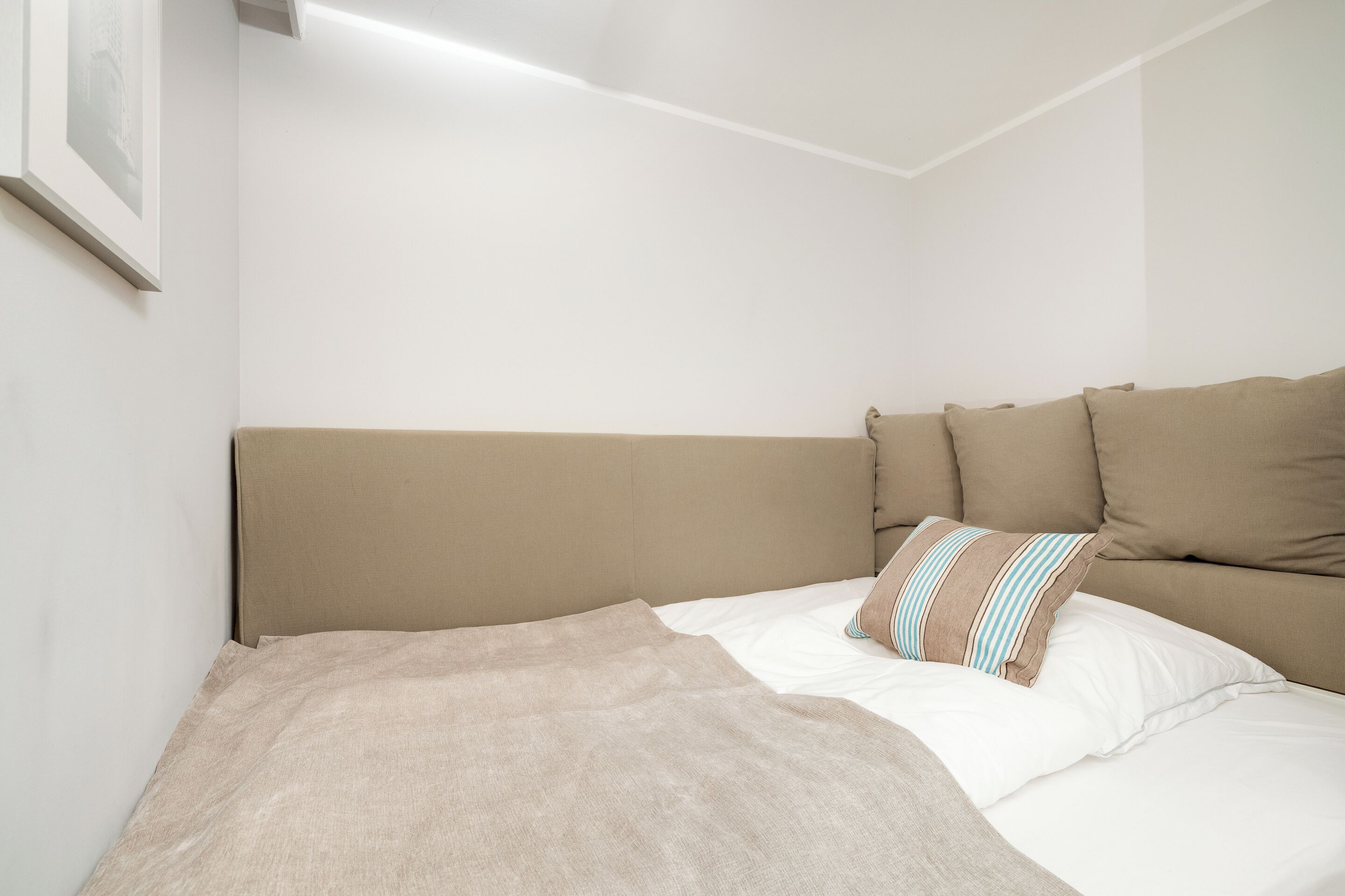 Un letto ben rifatto con una coperta beige e cuscini, una parete bianca liscia e un quadro appeso a sinistra.