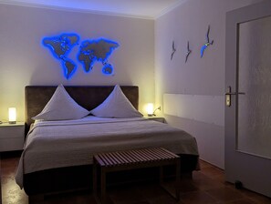 Una cama bien tendida con un mapa mundial iluminado en azul, un banco de madera y una puerta con tres relojes de pared.