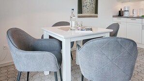 Una zona de menjador amb una taula blanca, quatre cadires grises tapissades, una ampolla, un got i un quadern. Una làmpada penjant hi penja i un vas decoratiu a la paret.