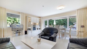 Een moderne woonkamer met een flatscreen televisie, een glazen salontafel, een zwarte leren bank en een eethoek met witte stoelen en een tafel.