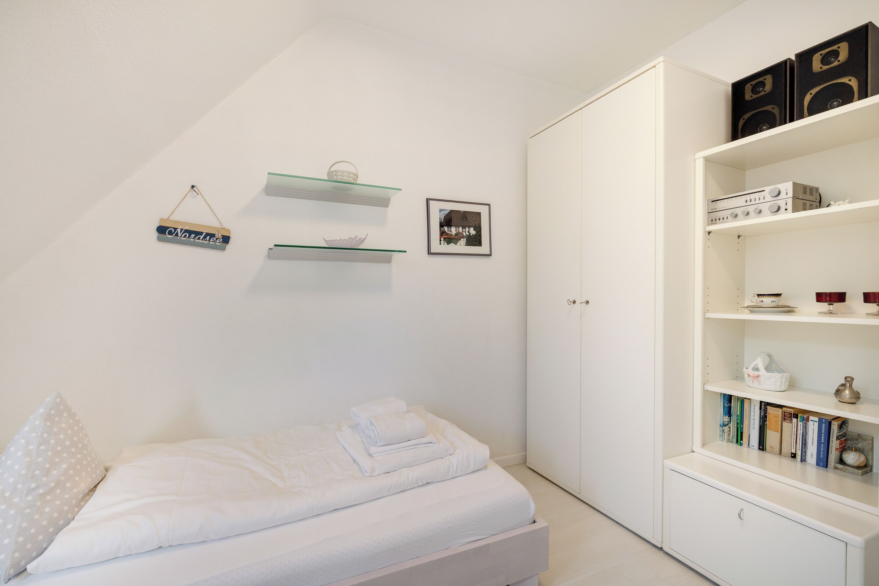 A bedroom with a bed, a wardrobe, a bookshelf, and a wall-mounted shelf with a picture and a sign.
