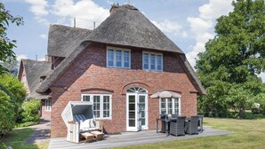 A brick house with a steeply pitched roof, a front porch with a wicker chair and table, and a well-maintained lawn.