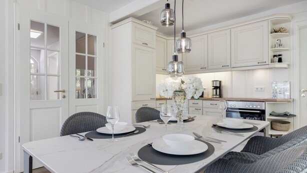 A modern kitchen with white cabinetry, a marble countertop, and a dining area set with white plates, glasses, and cutlery.