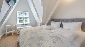 A bedroom with a sloped ceiling, a bed with white bedding, a grey upholstered headboard, and a window with blinds.