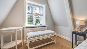 A room with a sloped ceiling, a window with blinds, a white bench, a small table, and a lamp.