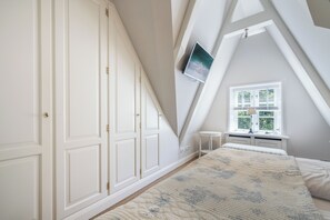 A bedroom with a sloped ceiling, a bed with a quilt, a window with blinds, and a wall-mounted TV. - Muschelstrand - Sehr Großzügig Geschnittene Obergeschoss-wohnung am Wattenmeer (Sylt / OT Keitum)