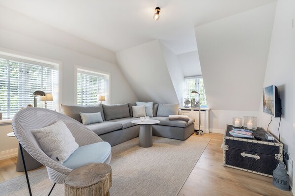 Ein modernes Wohnzimmer mit einem grauen Ecksofa, einem runden Couchtisch, einem Korbsessel und einem kleinen Beistelltisch mit Kerzen.