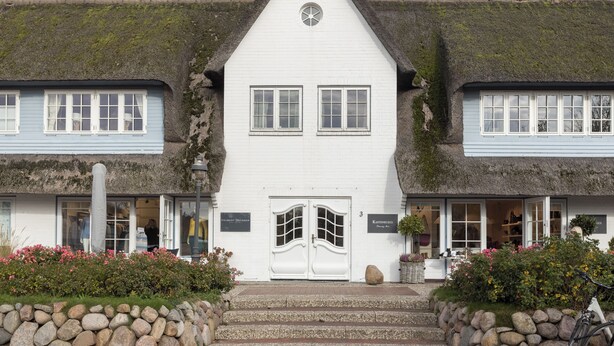 Un bâtiment traditionnel aux murs blanchis et doté d’un toit de chaume, avec une entrée centrale flanquée d’une double porte et une petite fenêtre circulaire au-dessus de la porte.