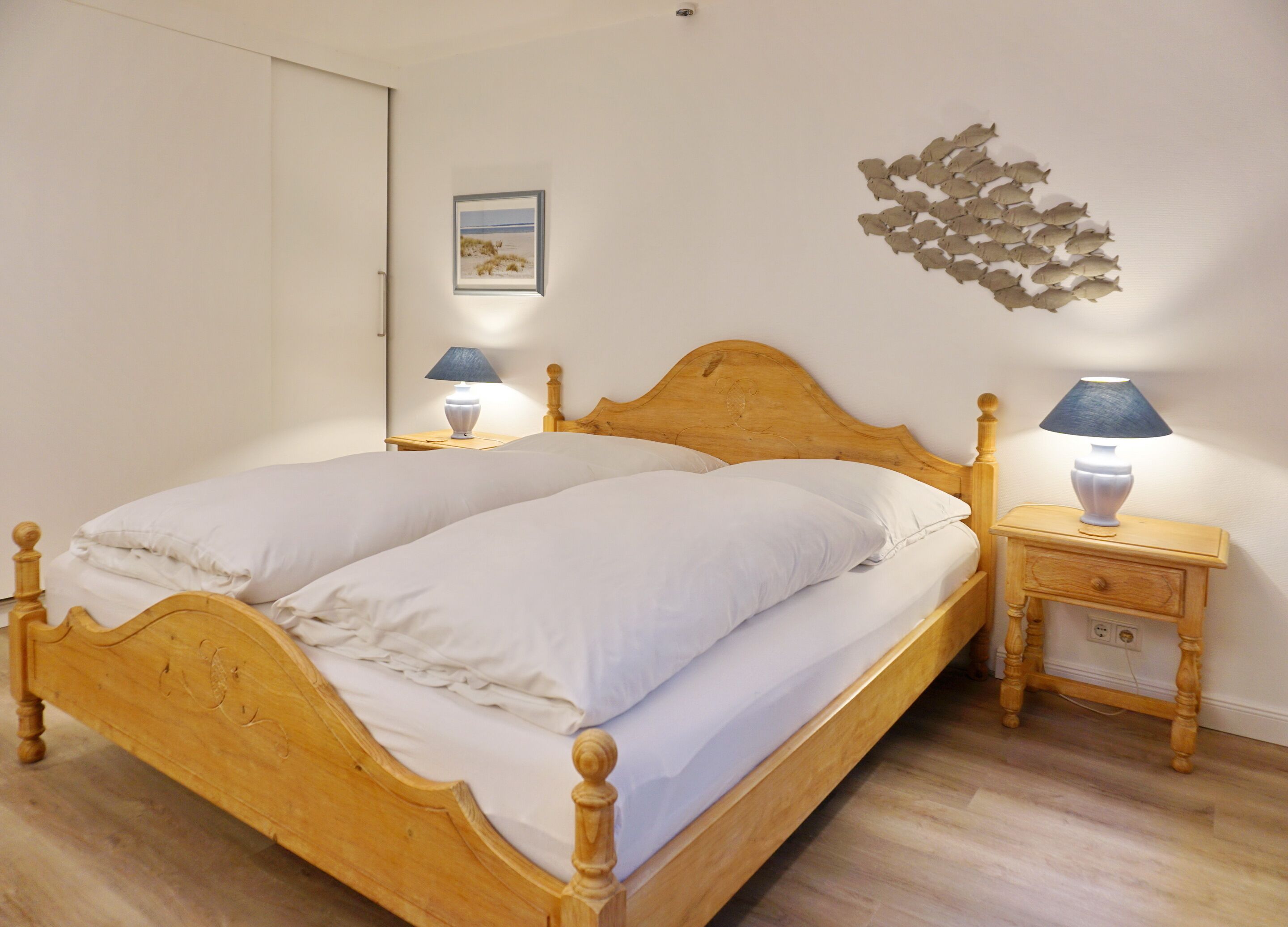 A double wooden bed with white linens, two bedside tables with lamps, a wall-mounted abstract art piece, and a framed picture above the bed.