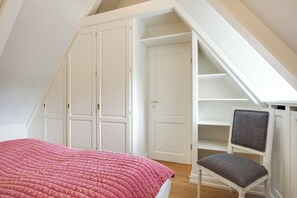 A bedroom with a sloped ceiling, a bed with a pink quilt, a chair with a grey cushion, and white wardrobes. - Großzügig Geschnittene Og-wohnung im Historischen Kern von Keitum (Sylt / OT Keitum)