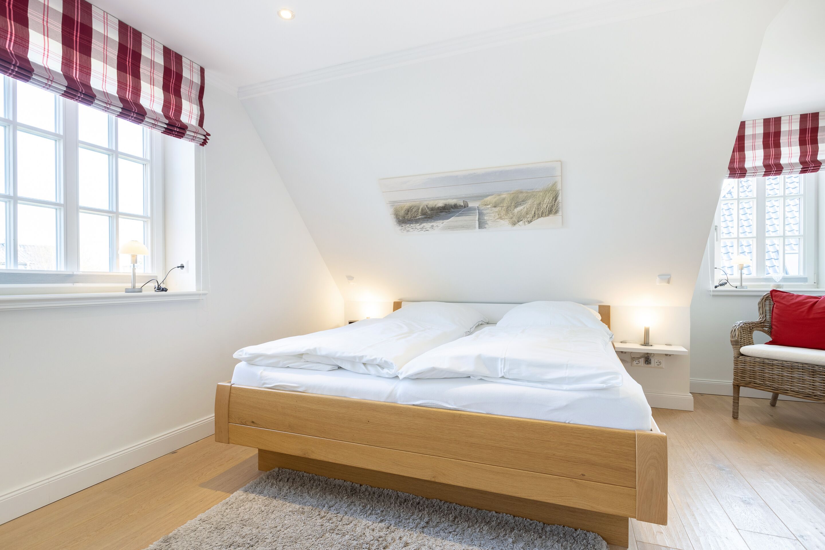 A bedroom with a wooden bed, white bedding, a painting on the wall, and a window with red curtains.