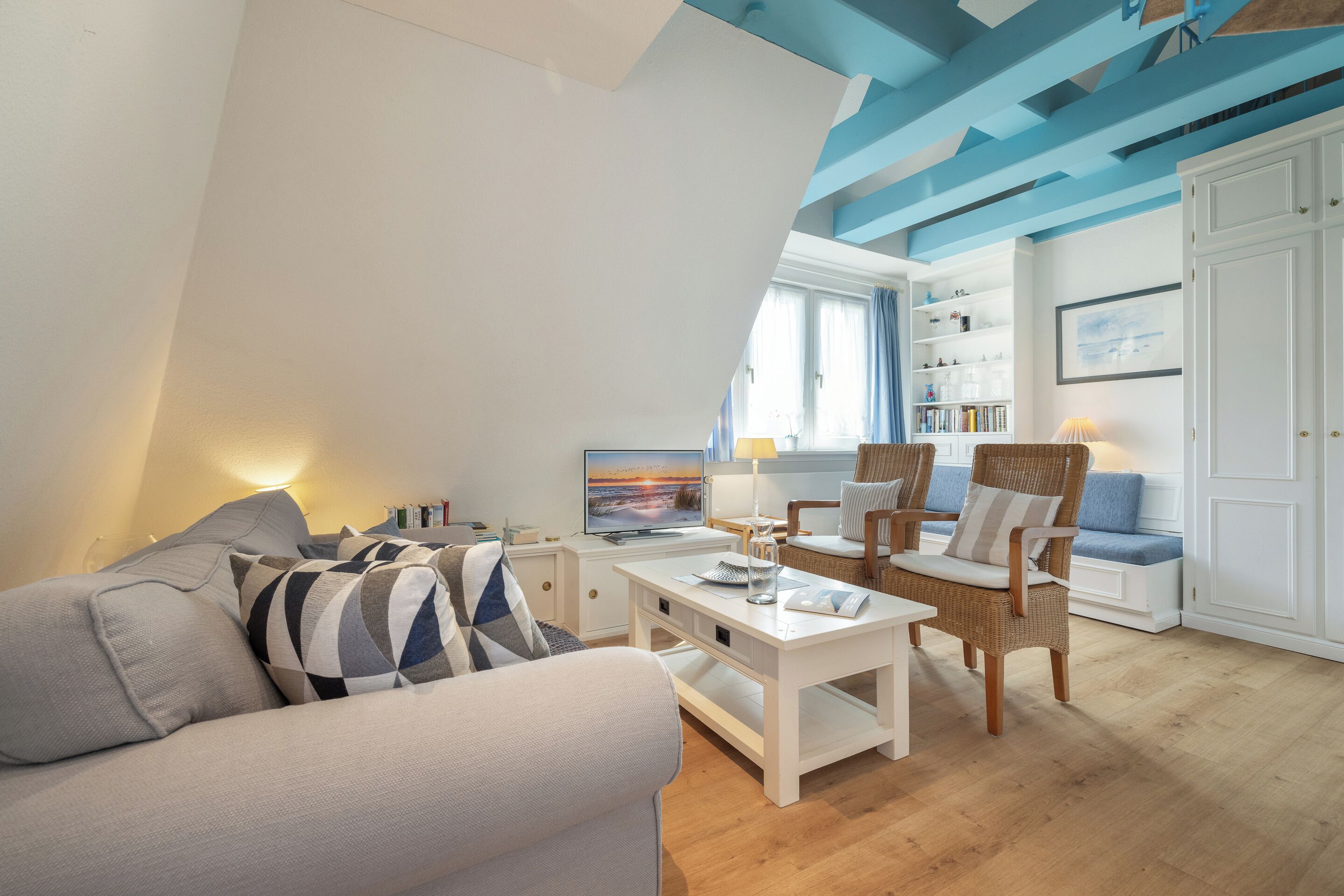 Ein modernes Wohnzimmer mit einer Couch, einem Couchtisch und einem Sessel. Im Hintergrund steht ein Bücherregal und ein Fernseher.