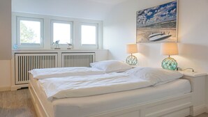 Ett sovrum med en stor säng, två sänglampor, ett fönster med persienner, ett element och en tavla med strandmotiv på väggen.